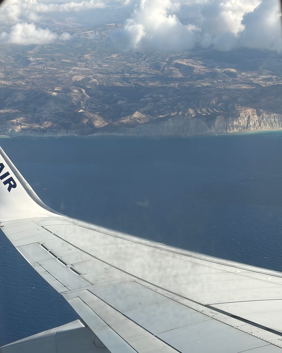 The Cyprus coast seen from a plane window, with the wing in front and clouds over the mountains